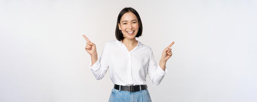 Image of beautiful asian woman pointing fingers left and right, making decision, showing two variants choices, standing over white background.