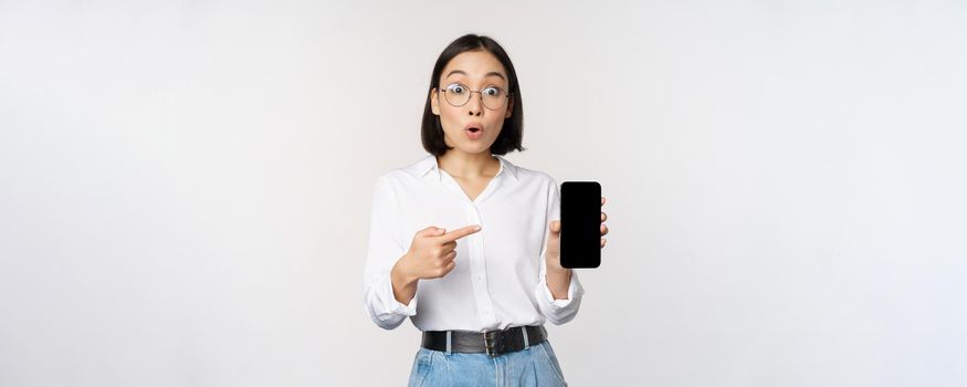 Enthusiastic young asian woman pointing finger at smartphone screen, showing advertisement on mobile phone, white background.