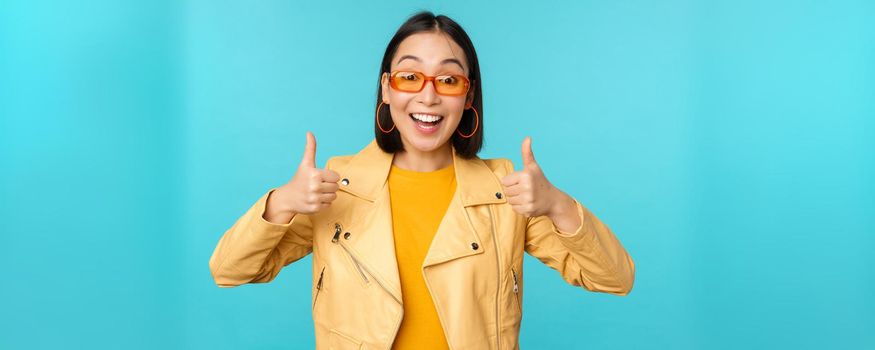 Enthusiastic korean girl looks excited and shows thumbs up, approves smth awesome, excellent choice and quality, stands over blue background.