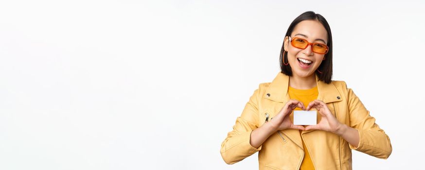 Stylish attractive asian girl in sunglasses, showing credit card and smiling, standing happy against white studio background. Copy space
