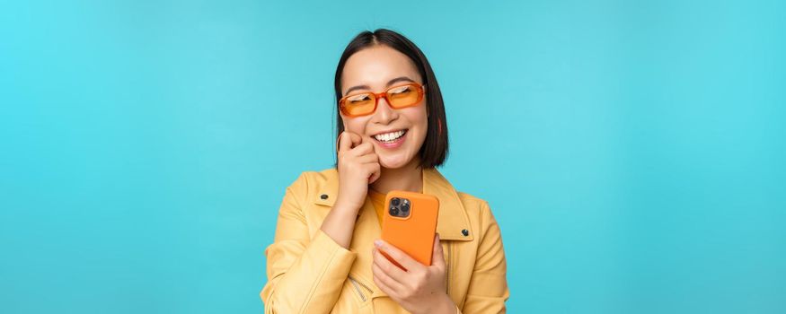 Happy stylish asian girl using smartphone and laughing, smiling at camera, standing over blue background. Copy space