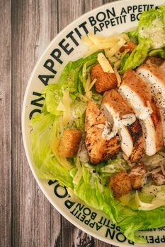 Caesar salad with thinly sliced chicken breast and traditional aioli on a plate that says bon apetite on a rustic table. Top view. Close-up detail. Vertical orientation.