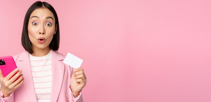 Amazed businesswoman, asian girl in suit showing credit card and mobile phone, order online, shopping with smartphone, standing over pink background. Advertising concept.