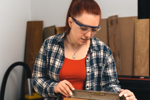 Close-up of the hands of a young red-haired carpenter, concentrated and precise, working on the layout of timbers in a small carpentry workshop, dressed in a blue checked shirt and a red T-shirt. Female carpenter using a precision level in her small carpentry workshop. Warm light indoors, background with wooden slats. Horizontal.