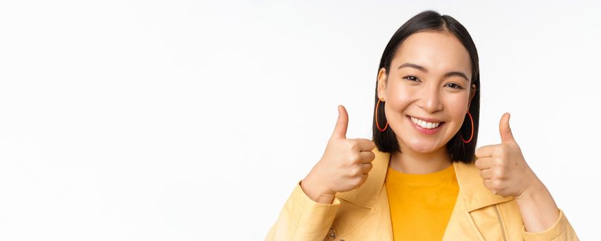 Smiling asian girl showing thumbs up, looking pleased, approve smth, standing over white background. Copy space