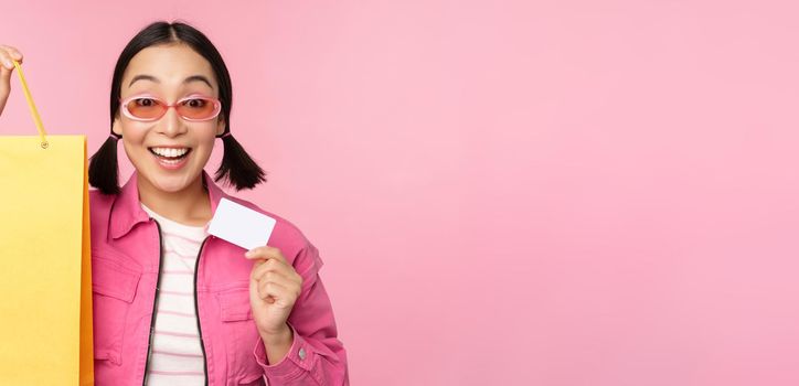 Happy young asian woman showing credit card for shopping, holding bag, buying on sale, going to the shop, store, standing over pink background.
