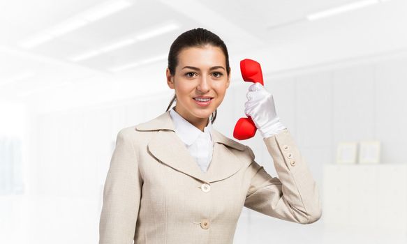 Smiling young woman holding retro red phone. Call center operator in white business suit posing with telephone in light office interior. Hotline telemarketing. Business assistance and consultation.