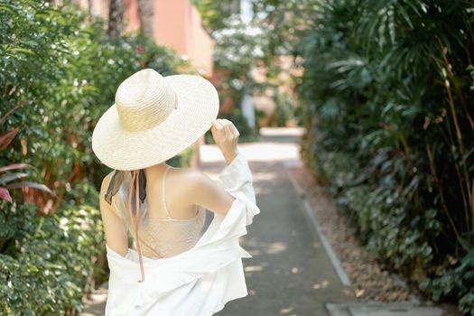 Back view of woman wearing white shirt, long skirt and straw hat posing tropical garden.