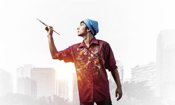 Young emotional artist gesturing with paintbrush. Happy painter in shirt and bandana standing on background modern office buildings. Creative hobby and artistic occupation concept with copy space