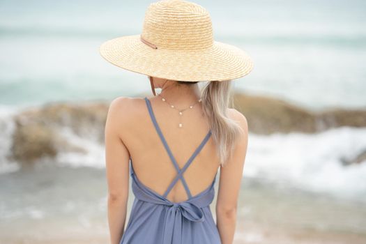 Back side of woman in blue dress and straw hat, standing on a timber by the ocean.