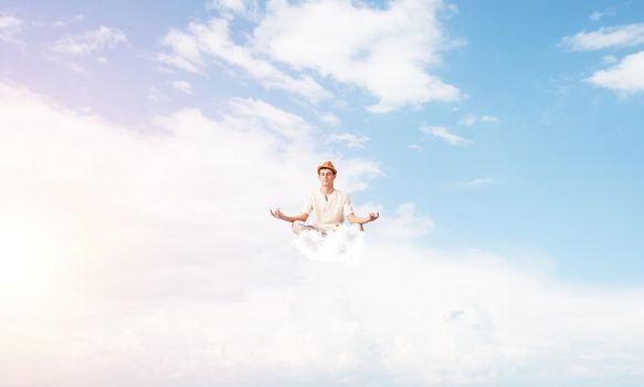 Man in white clothing keeping eyes closed and looking concentrated while meditating on clouds in the air with cloudy skyscape on background.