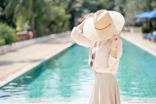 Woman wearing white shirt, long skirt and straw hat posing near swimming pool.