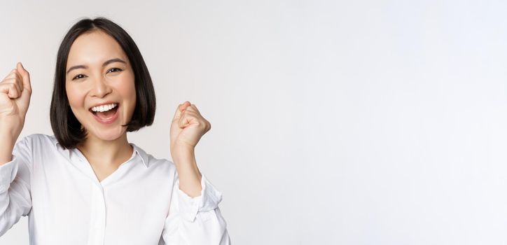 Close up face portrait of dancing asian woman smiling, triumphing and celebrating with happy emotion, standing over white background. Copy space