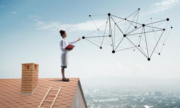 Horizontal shot of confident woman doctor in white medical suit standing at the top of brick building and interracting with network structure. Modern medical industry concept