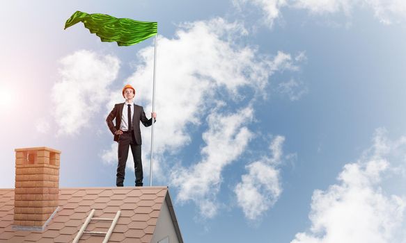 Businessman standing on house roof and holding green flag. Mixed media