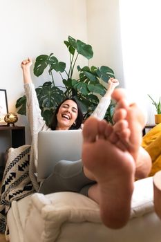 Excited young caucasian woman raising arms up celebrating success or achievement at home using laptop. Copy space. Vertical image. Victory concept.