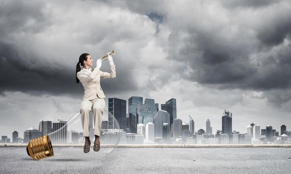 Attractive woman playing trumpet brass on background modern city and storming sky. Young businesslady in white suit sitting on big light bulb with music instrument. Musician practicing and performing