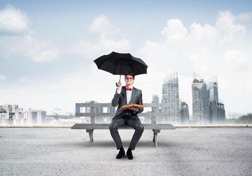 Young student with a book and umbrella sits on a wooden bench