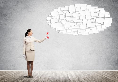 Young woman holding red phone and speech bubbles on grey wall background. Call center operator in white business suit. Business conversation and internet chatting. Professional business assistance