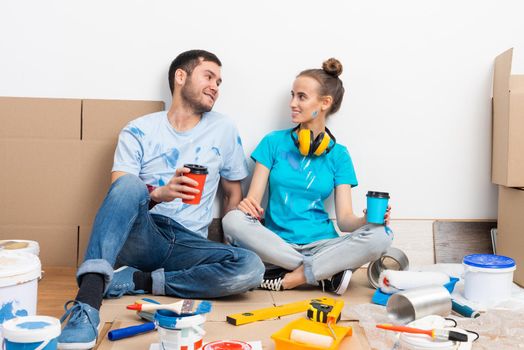 Happy boy and girl drinking coffee on floor. Home remodeling after moving. Cardboard boxes, construction tools and materials for building on floor. Couple having fun in time of house renovation.
