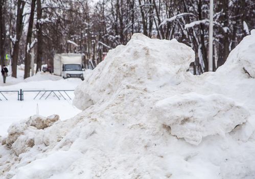 A high large snowdrift by the road against the backdrop of a city street. On the road lies dirty snow in high heaps. Urban winter landscape. Cloudy winter day, soft light.