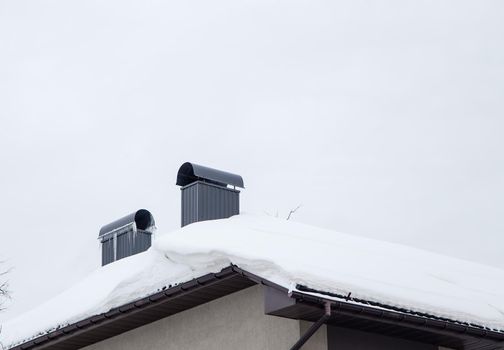 Two rectangular metal chimneys on the roof of the house. There is white snow on the surface. Against the background of a gray sky. Cloudy winter day, soft light.