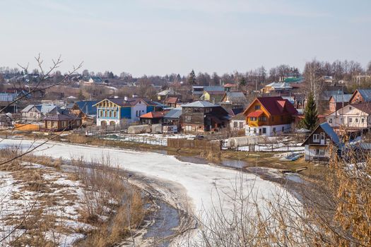 A frozen river stretching into the distance, a village on the bank. Spring, snow melts, puddles and dry grass all around. Day, cloudy weather, soft warm light.