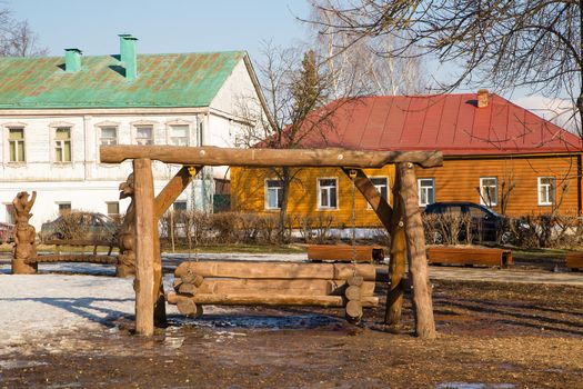 Wooden swing in the park against the backdrop of village houses. Spring, snow melts, dry grass all around, small snowdrifts and puddles. Day, cloudy weather, soft warm light.