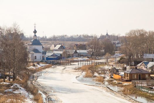 A frozen river stretching into the distance, a bridge in the distance. Spring, snow melts, puddles and dry grass all around. Day, cloudy weather, soft warm light.