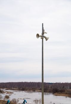 Two loudspeakers-megaphones on a high pole against the background of the river. Spring, the snow is melting, there are puddles of slush and mud all around. Day, cloudy weather, soft warm light.