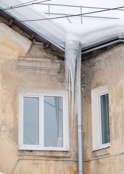 Dangerous ice icicles hang from the drainpipe at the edge of the roof. Against the background of the wall of an old brick house. Large cascades, even beautiful rows. Cloudy winter day, soft light.