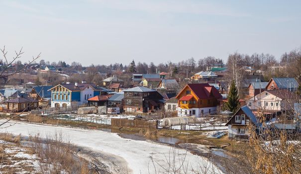 A curved, frozen river stretching into the distance, wooden houses on the shore. Spring, snow melts, puddles and dry grass all around. Day, cloudy weather, soft warm light.