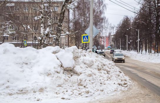 A huge large snowdrift by the road against the backdrop of a city street. On the road lies dirty snow in high heaps. Urban winter landscape. Cloudy winter day, soft light.