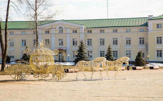 Outdoor sculpture of a horse with a carriage on a frame of light bulbs. Spring, snow melts, in the background is a wall of a multi-storey building with windows. Day, cloudy weather, soft warm light.