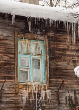 Glass transparent icicles hang on the edge of the roof and the window. Against the background of the wooden wall of the old house. Large cascades, even beautiful rows. Cloudy winter day, soft light.