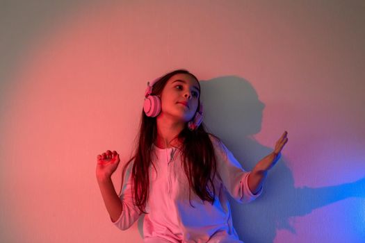 A dancing happy little girl in pink headphones with loose dark hair, in neon pink blue light near the wall