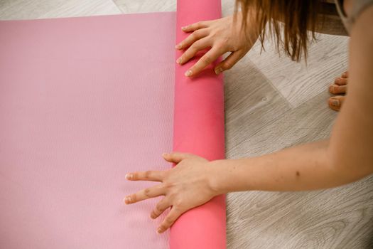 A young woman rolls a pink fitness or yoga mat before or after exercising, exercising at home in the living room or in a yoga studio. Healthy habits, keep fit, weight loss concept. Closeup photo.