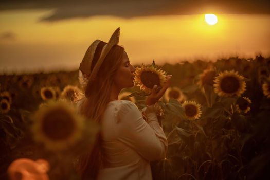 Beautiful middle aged woman looks good in a hat enjoying nature in a field of sunflowers at sunset. Summer. Attractive brunette with long healthy hair