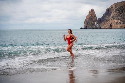 A beautiful and sexy brunette in a red swimsuit on a pebble beach, Running along the shore in the foam of the waves.