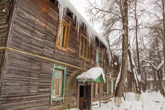 Growing transparent icicles hang on the edge of the roof. Against the background of the wooden wall of the old house. Large cascades, even beautiful rows. Cloudy winter day, soft light.