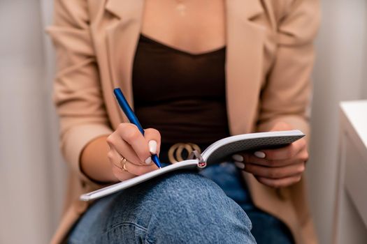 European woman writes in a notebook. She is wearing a beige jacket and jeans