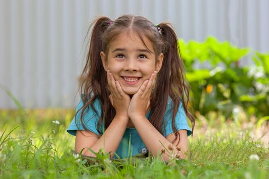 Adorable smiling brunette girl with two tails lies on the green grass, on her stomach, in the summer garden, holding two thumbs up