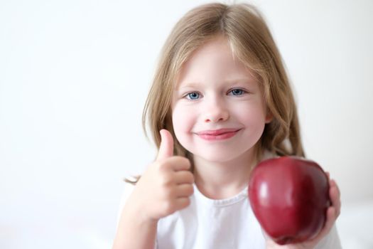 Little girl holding red apple in her hands and showing thumbs up. Proper balanced baby food concept