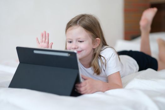 Little girl lying on bed and waving her hand at screen of tablet. Kids apps mobile concept