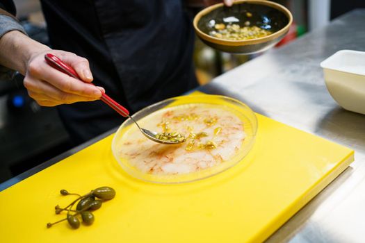 Anonymous cook spreading pine nut sauce on a plate of prawn carpaccio with capers while working in a restaurant kitchen.