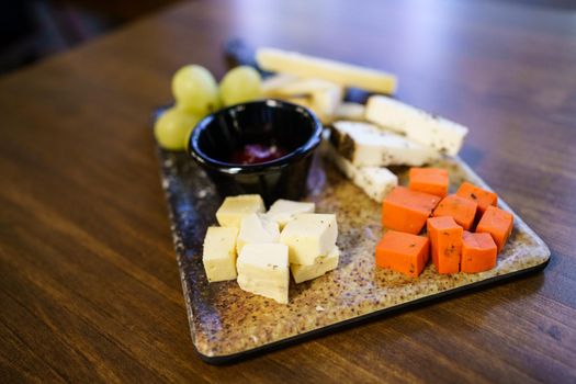 Ripe grapes and pieces of assorted cheese placed on board around bowl with sauce on restaurant table