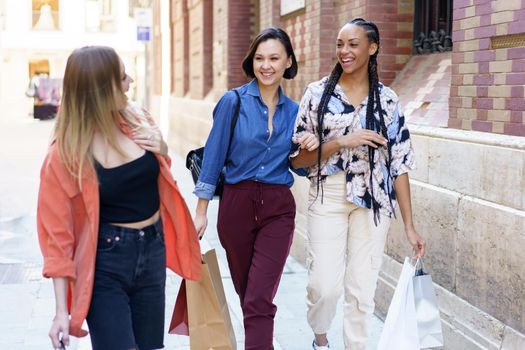 Cheerful multiracial female friends with shopping bags looking at each other while strolling together on walkway near building in city