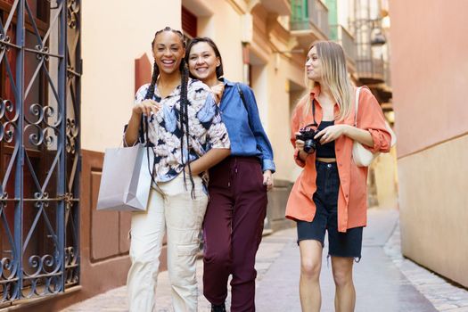Smiling fair haired woman with camera looking at cheerful diverse female friends while going home after buying new clothes