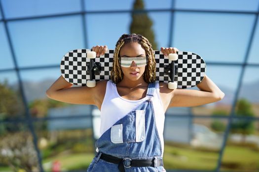 Black young woman dressed casual, looking at camera with modern sunglasses and a skateboard.