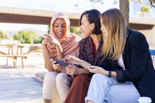 Content young multiracial female best friends, in casual clothes and hijab, smiling while watching video on smartphone sitting on bench in city park on sunny day